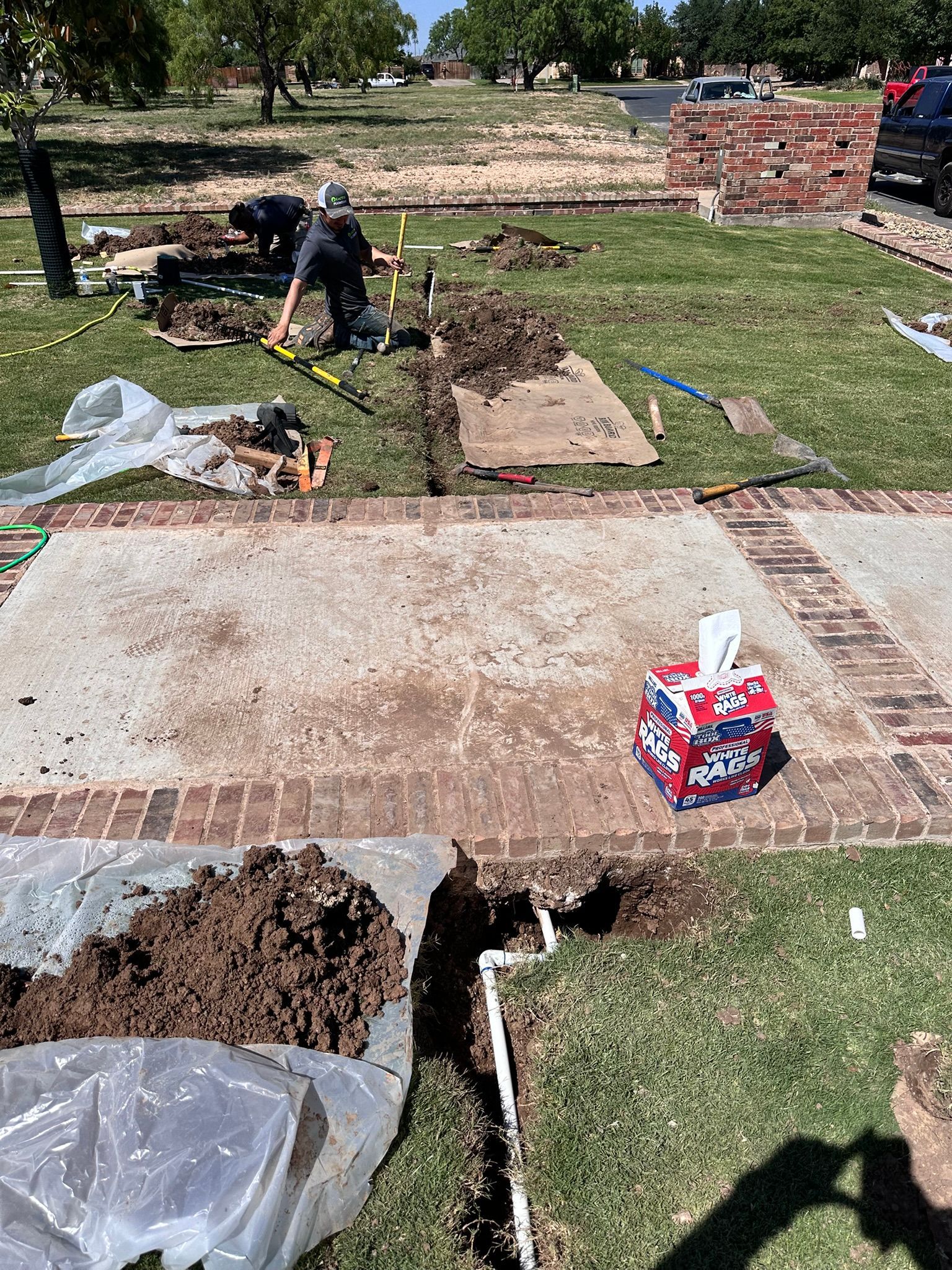 Workers digging trenches in a grassy yard, likely for plumbing or utilities, sunny day, dirt piles.