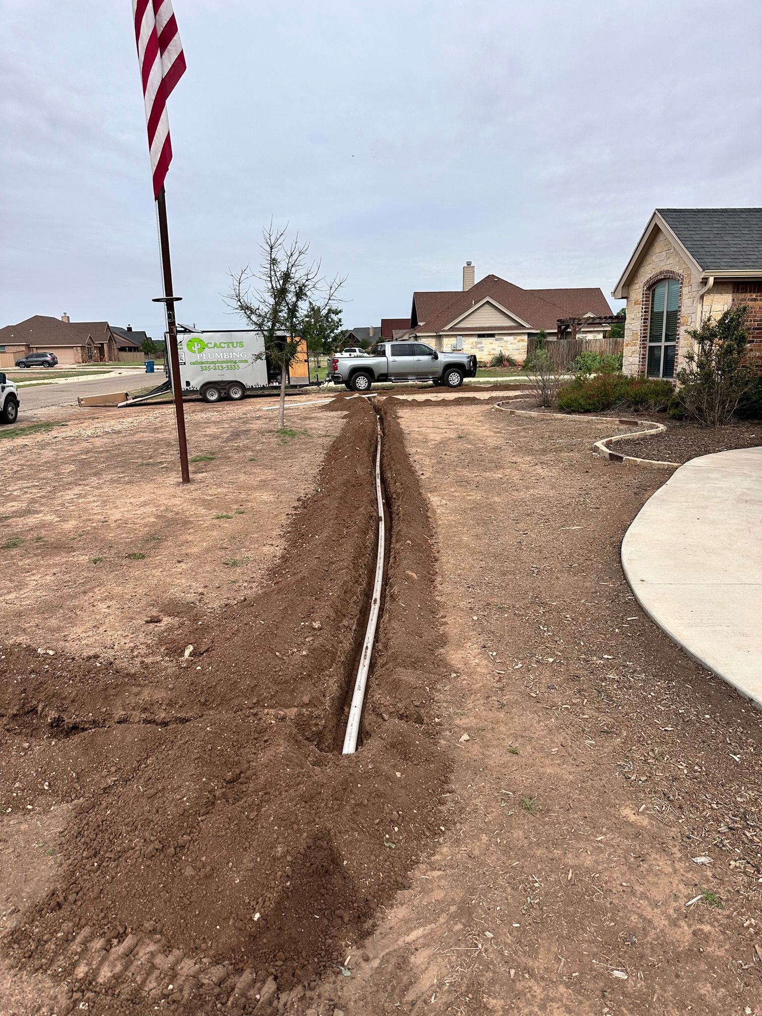 A trench dug in a yard with white pipe, flag, truck, and homes in the background.