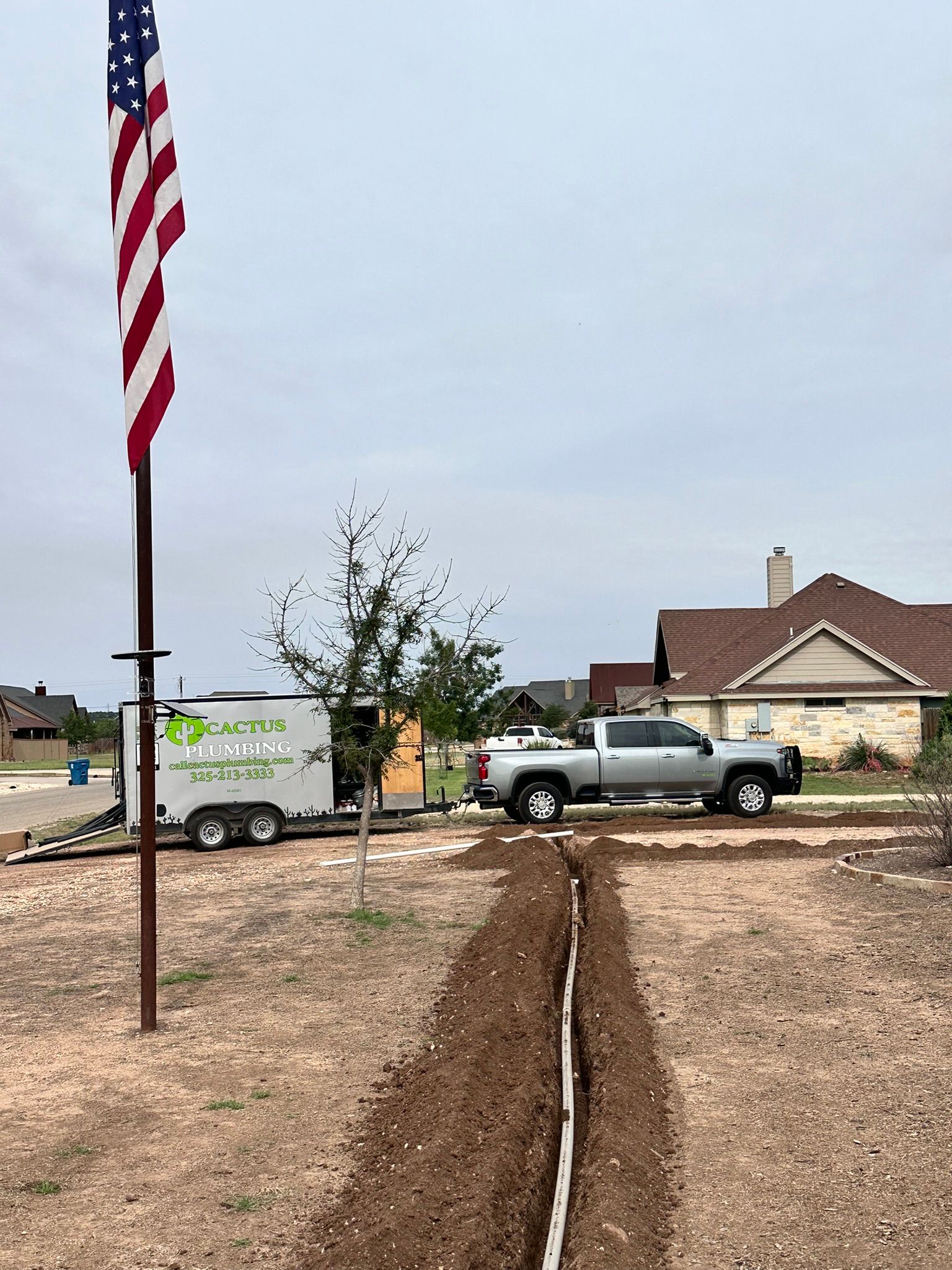 A trench with a white pipe, truck, trailer, and US flag in a yard.
