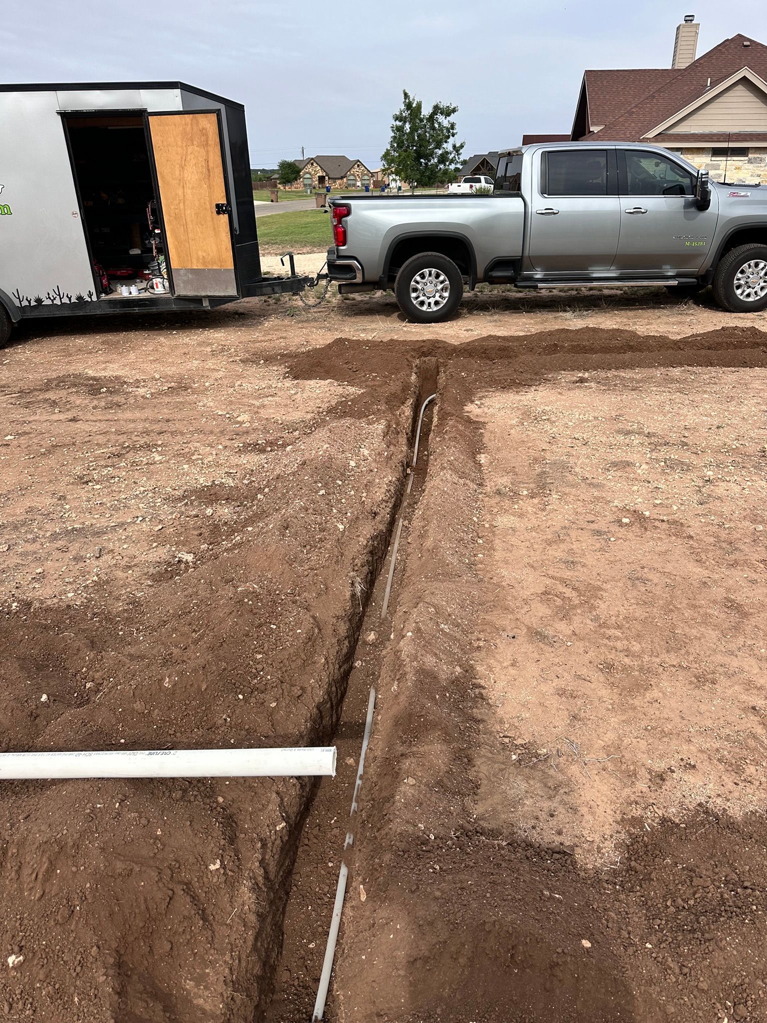 Trench dug in dirt with white pipes laid, near a truck and trailer in a residential area.