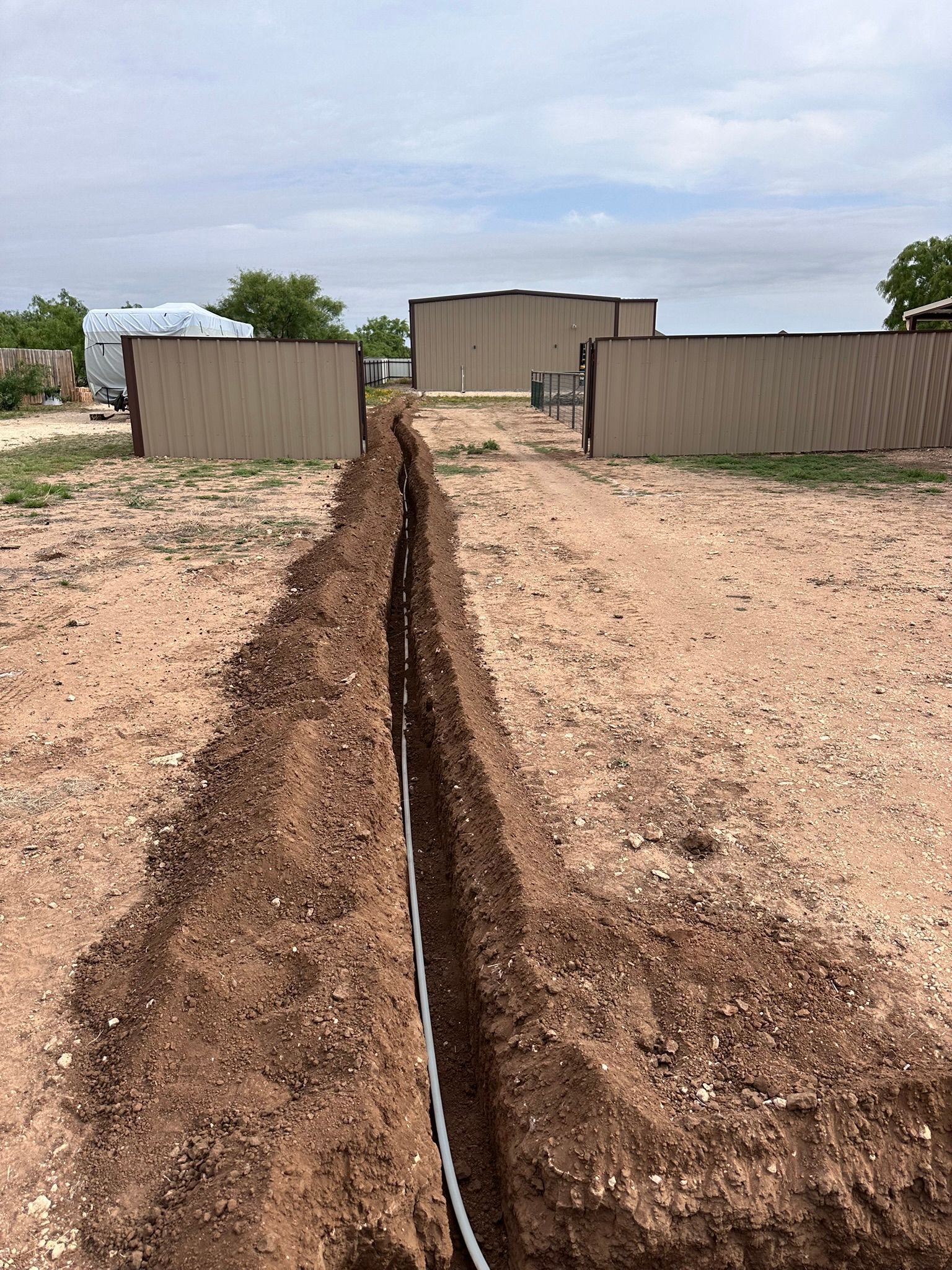 Trench with white pipe running through it in a dirt yard, with buildings in the background under a cloudy sky.