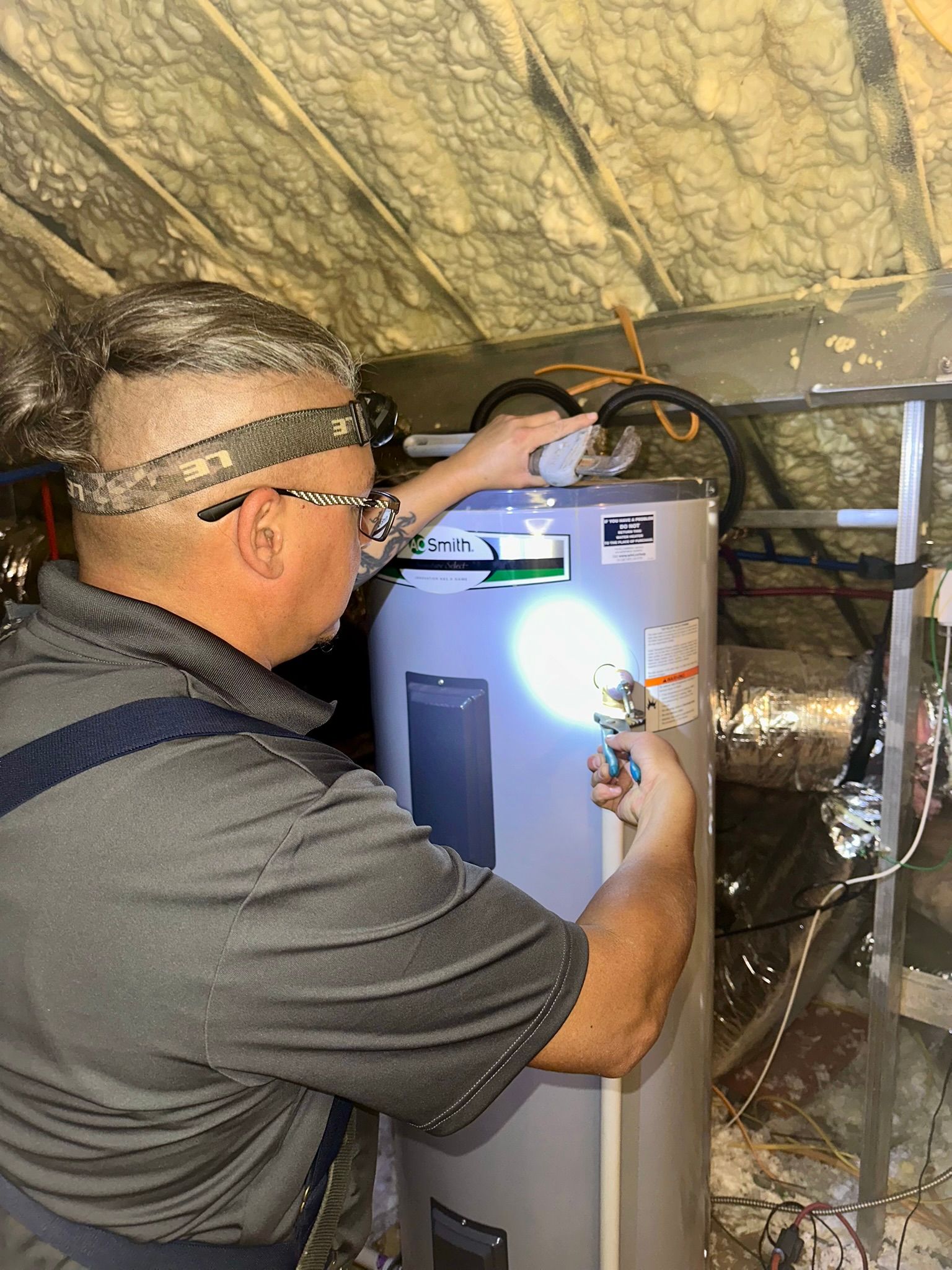 Man with headlamp examines water heater in attic.