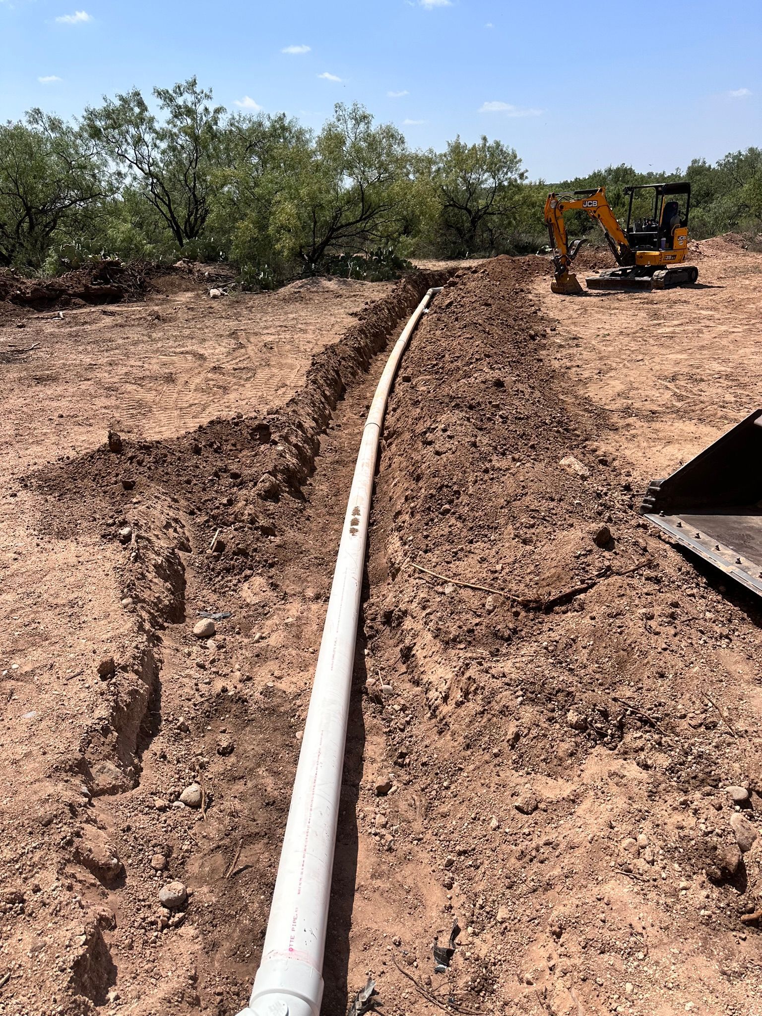 A white pipe laid in a narrow trench. A small excavator sits in the background; brown dirt and trees.