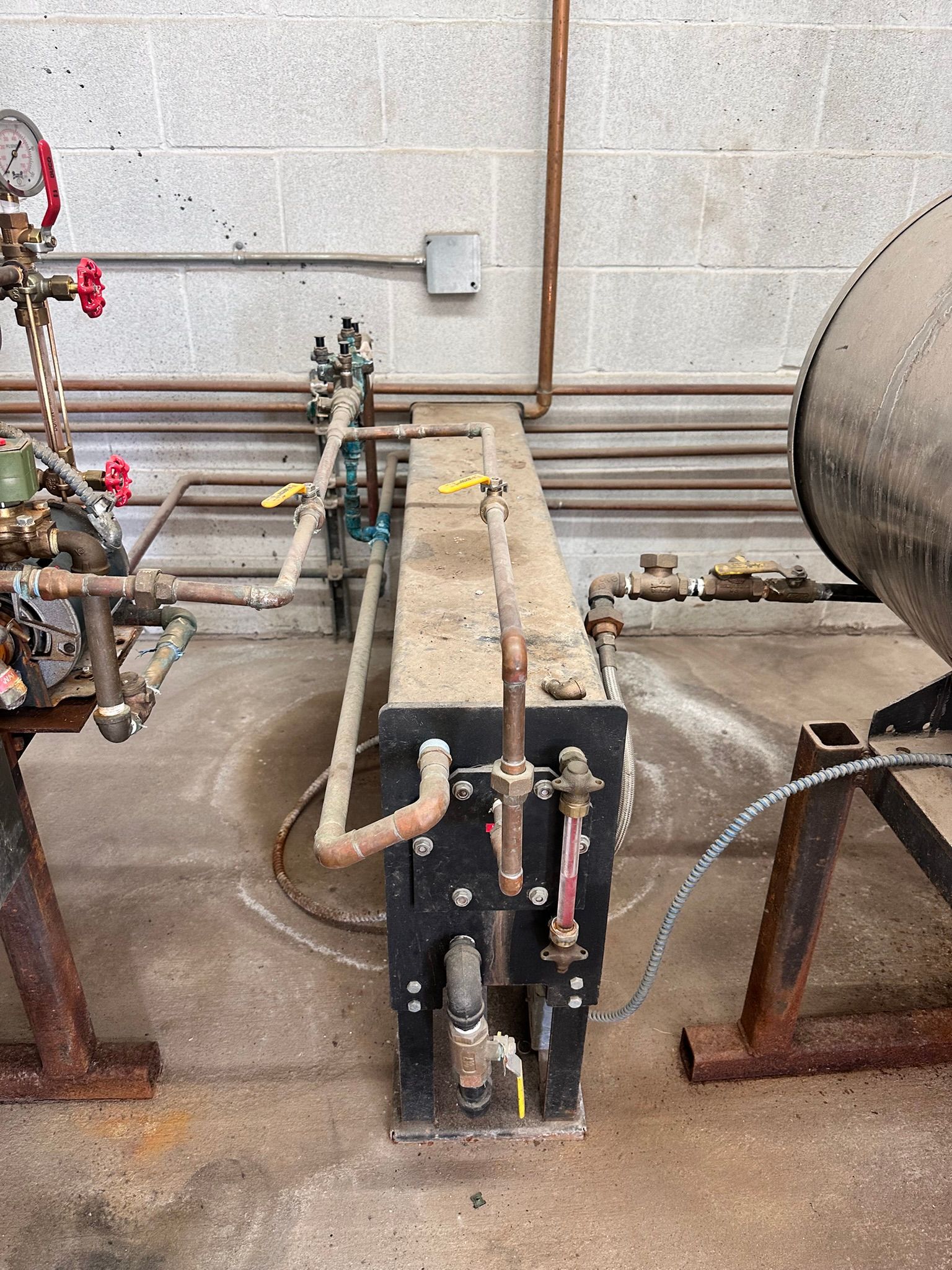 Industrial plumbing system with pipes, valves, and a black rectangular tank against a cinder block wall.
