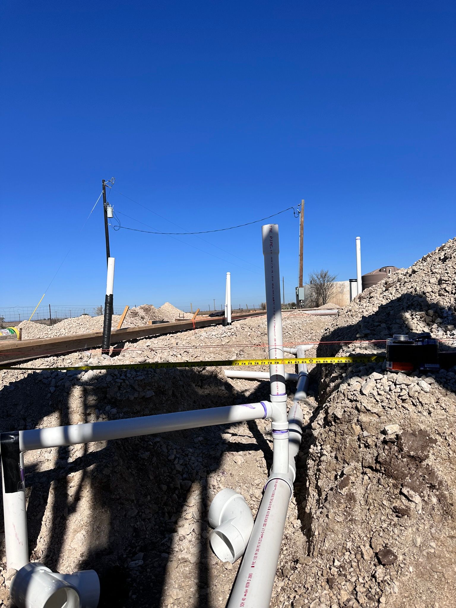 Construction site with white PVC pipes in foreground, piles of gravel, and blue sky.