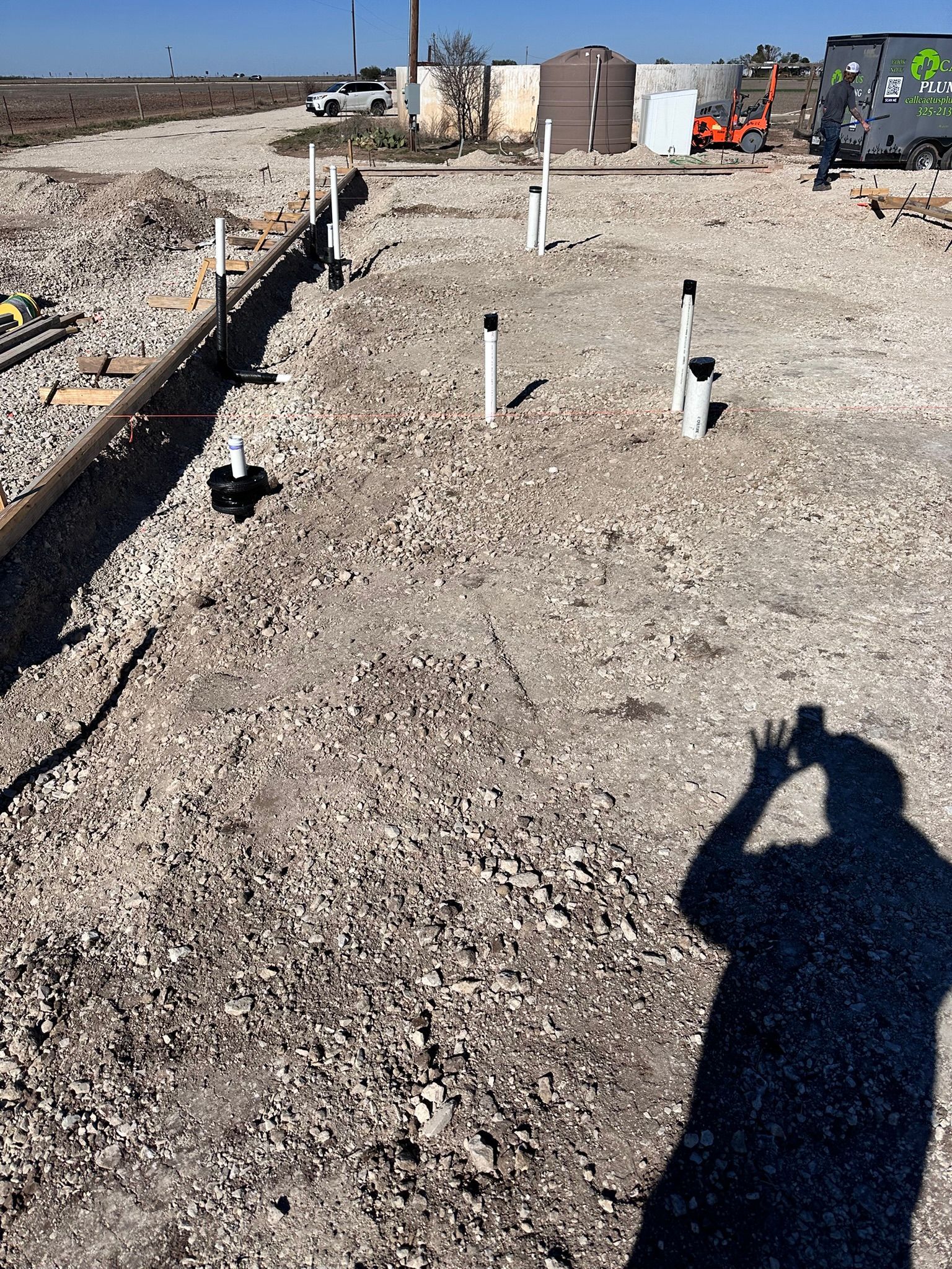 Construction site with exposed pipes, gravel ground, and person's shadow.