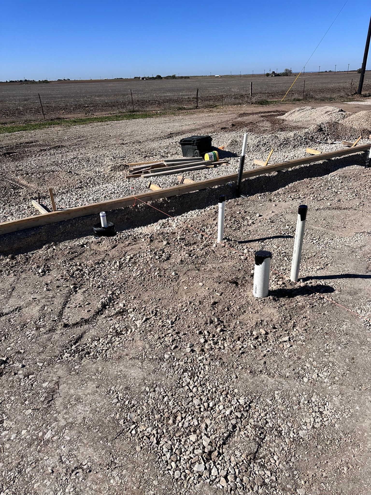 Gravel-covered construction site with PVC pipes, a long wooden beam, and a black box, under a clear blue sky.