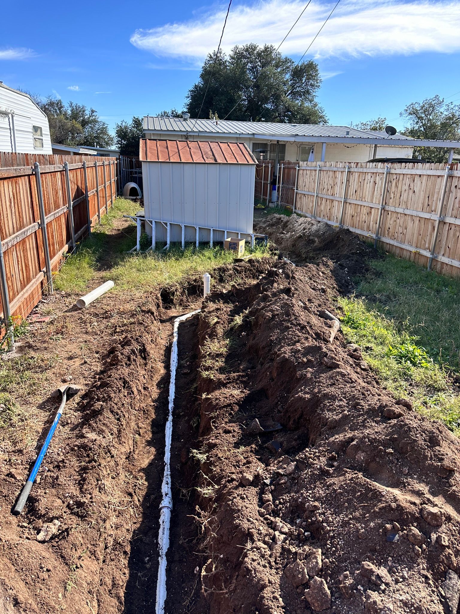 A backyard trench dug with white pipes, fences, shed, shovel, and blue sky.