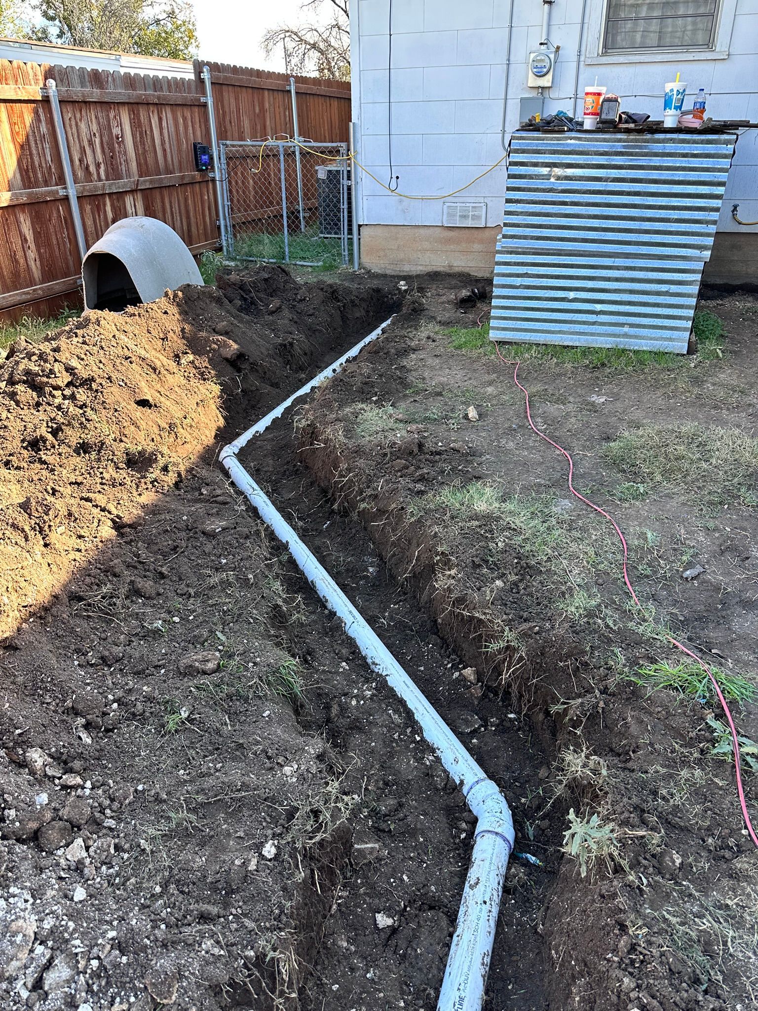 A trench with a grey pipe snaking through the yard, near a fence and corrugated metal shed.