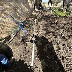 A trench dug along a brick wall, with tools, a white pipe, and a person working.
