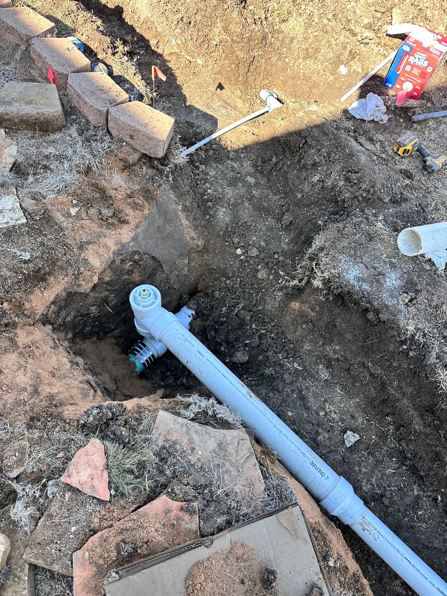 A gray PVC pipe and fittings installed in a trench, surrounded by soil and brick.