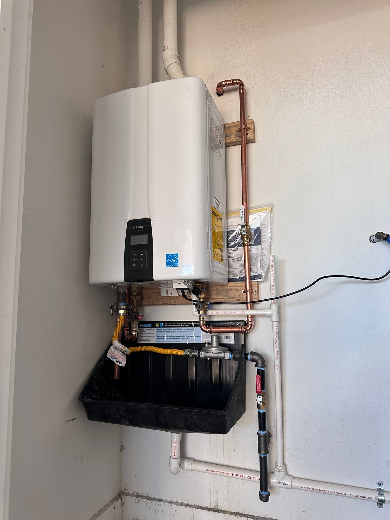 Tankless water heater mounted on a wall with copper pipes and black drain pan.