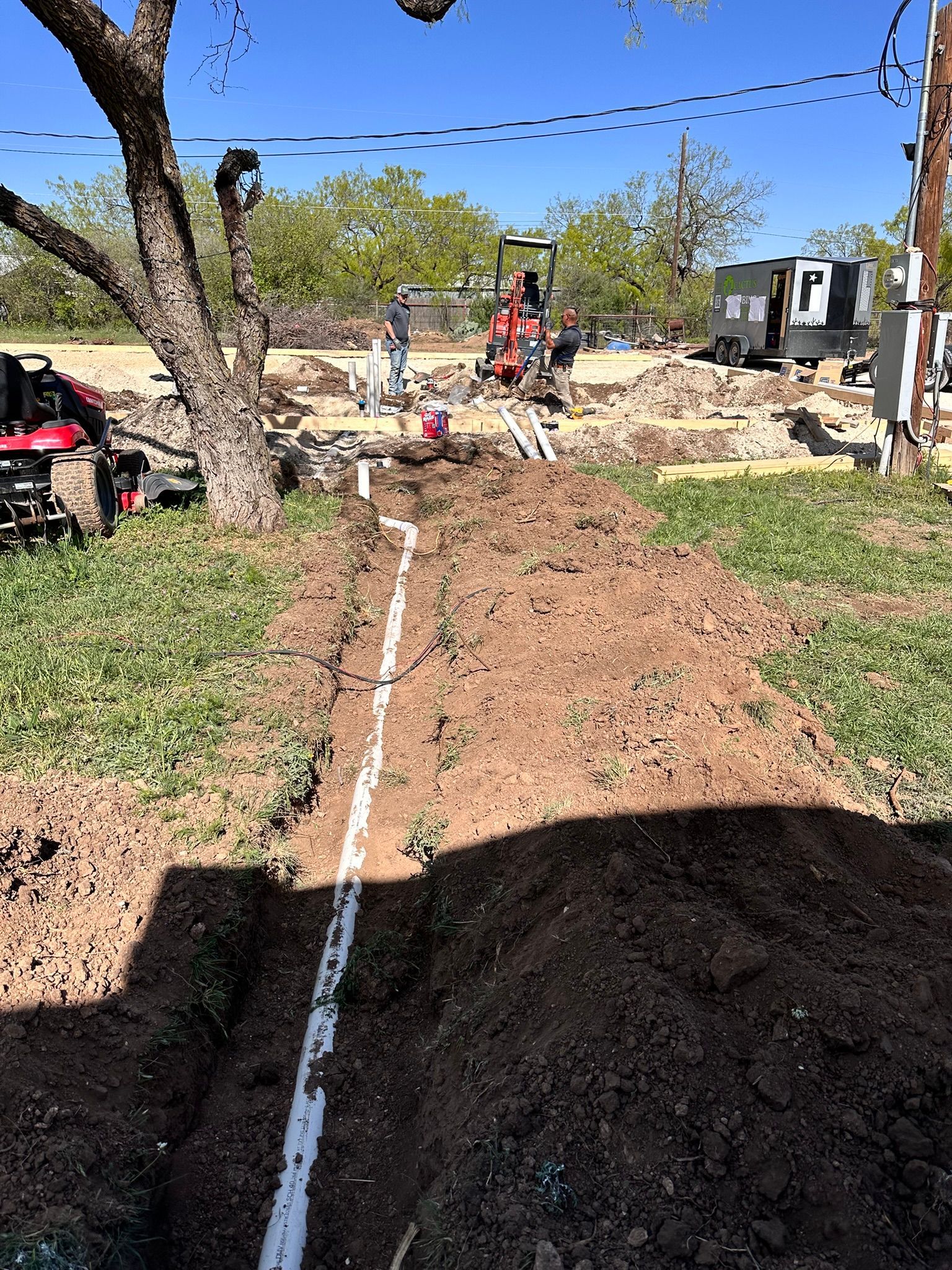 Trench dug in dirt, with white pipes laid. Workers and equipment in the background.