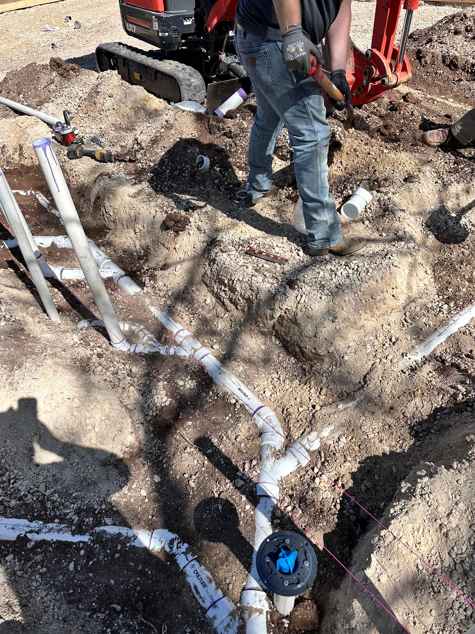 Person installing PVC pipes in a dirt trench with an excavator in the background.