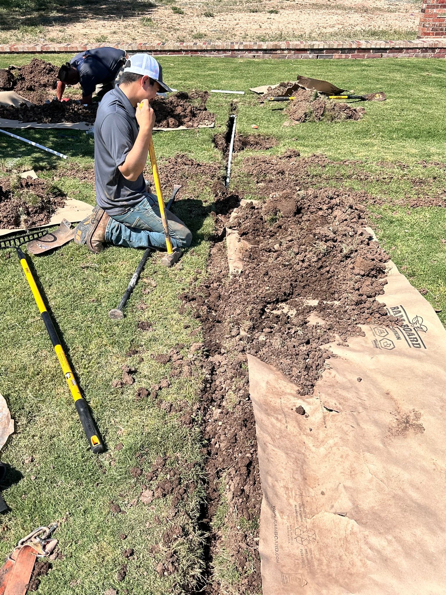 Two men digging in a grassy yard. One kneels with a shovel, the other works in the background.