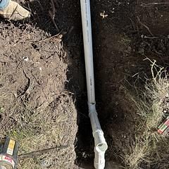 White PVC pipe installed in a trench in the ground, surrounded by soil and grass.