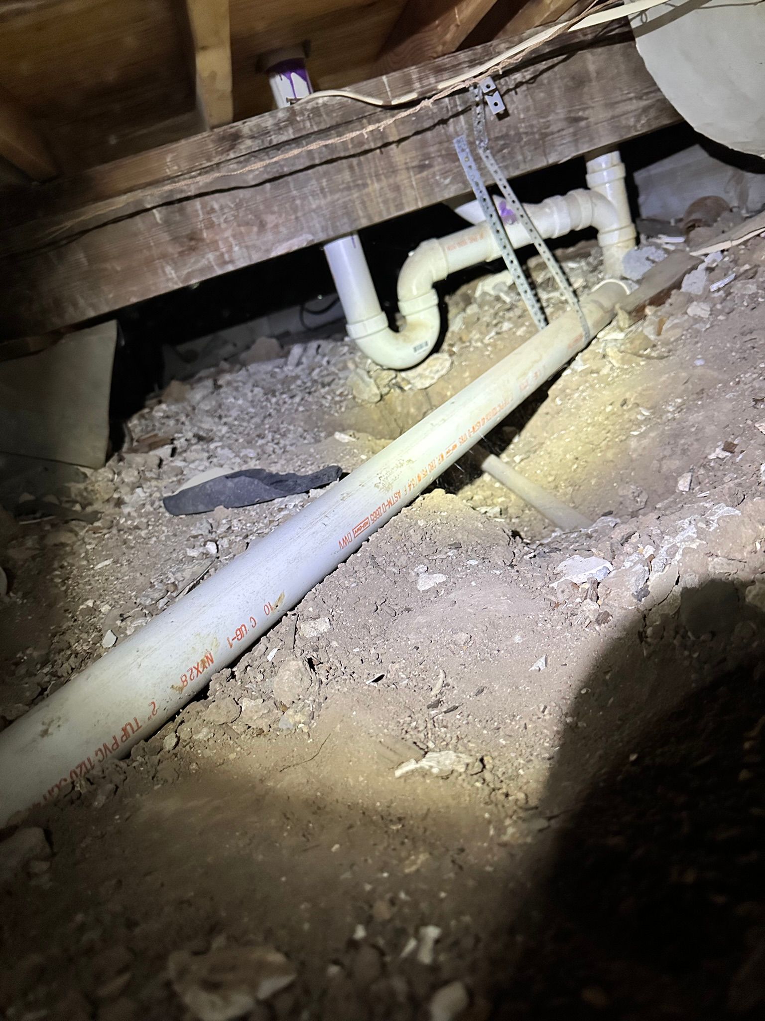Under-house view of plumbing pipes, dirt, and wood beams; white pipes connect to drainage system above.