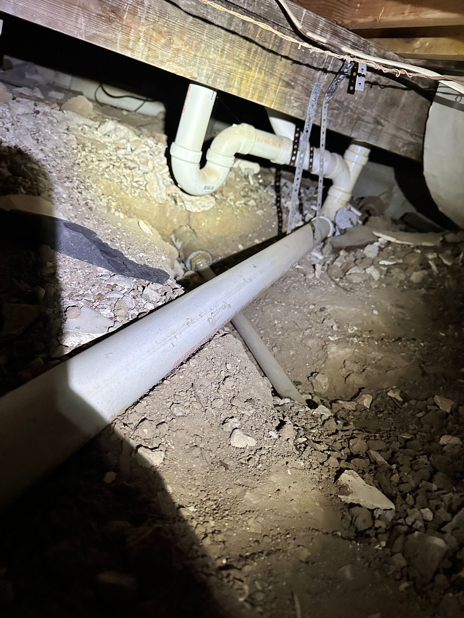 Underneath a building, PVC pipes wind through a dusty crawl space, alongside wood beams and debris.