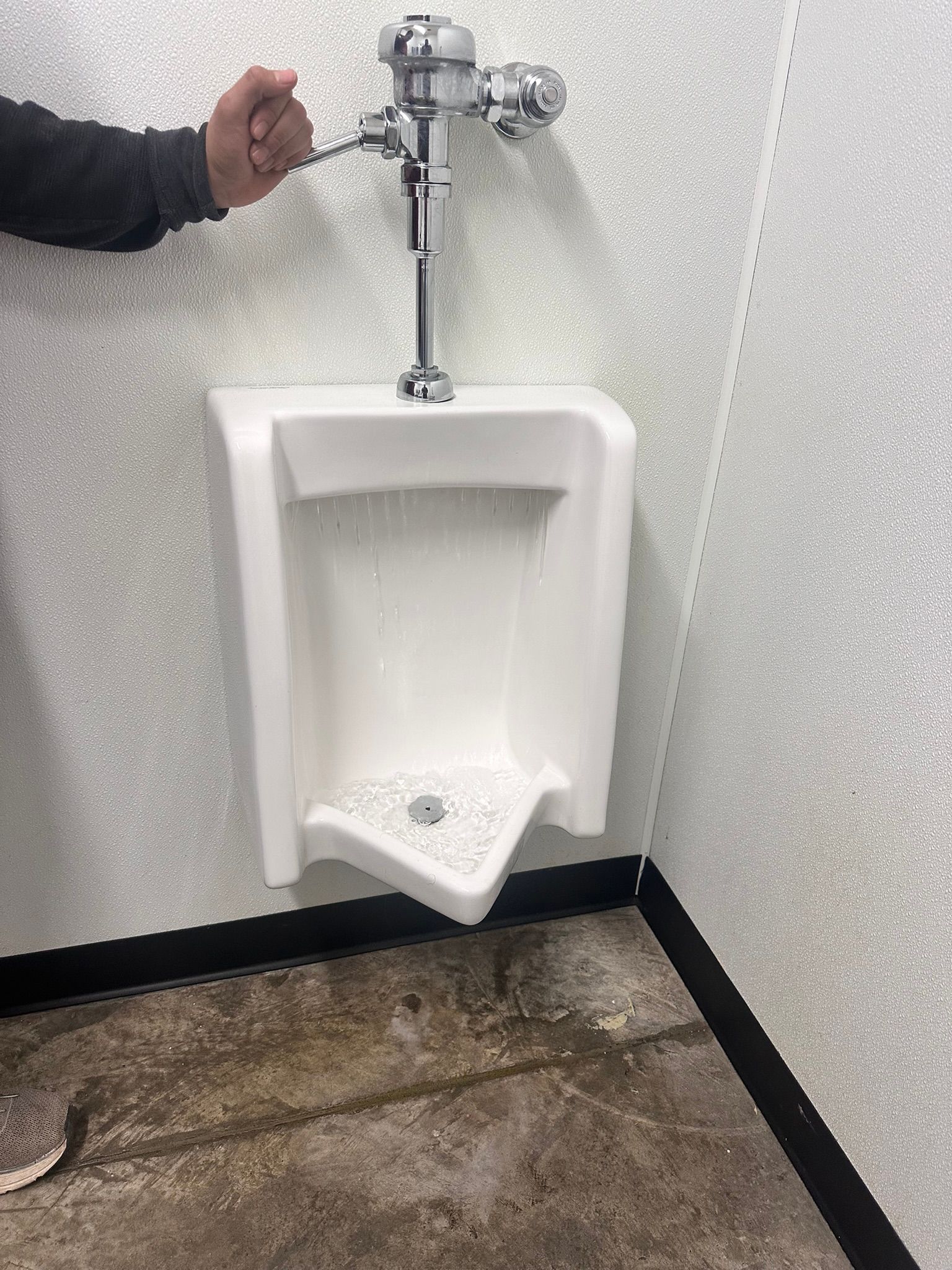 A hand operates the flush valve on a white urinal. The urinal is in a public restroom with white speckled walls.