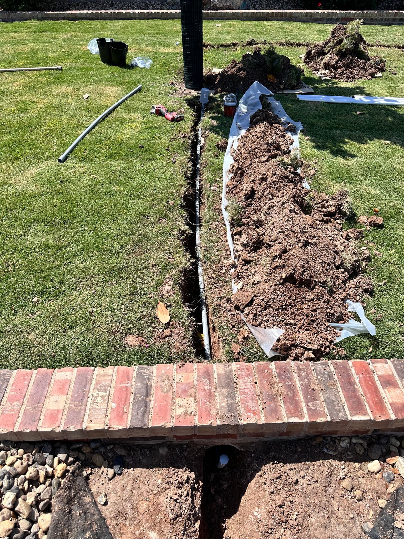 Trench dug in green grass, filled with dirt, near a brick border and utility pole.
