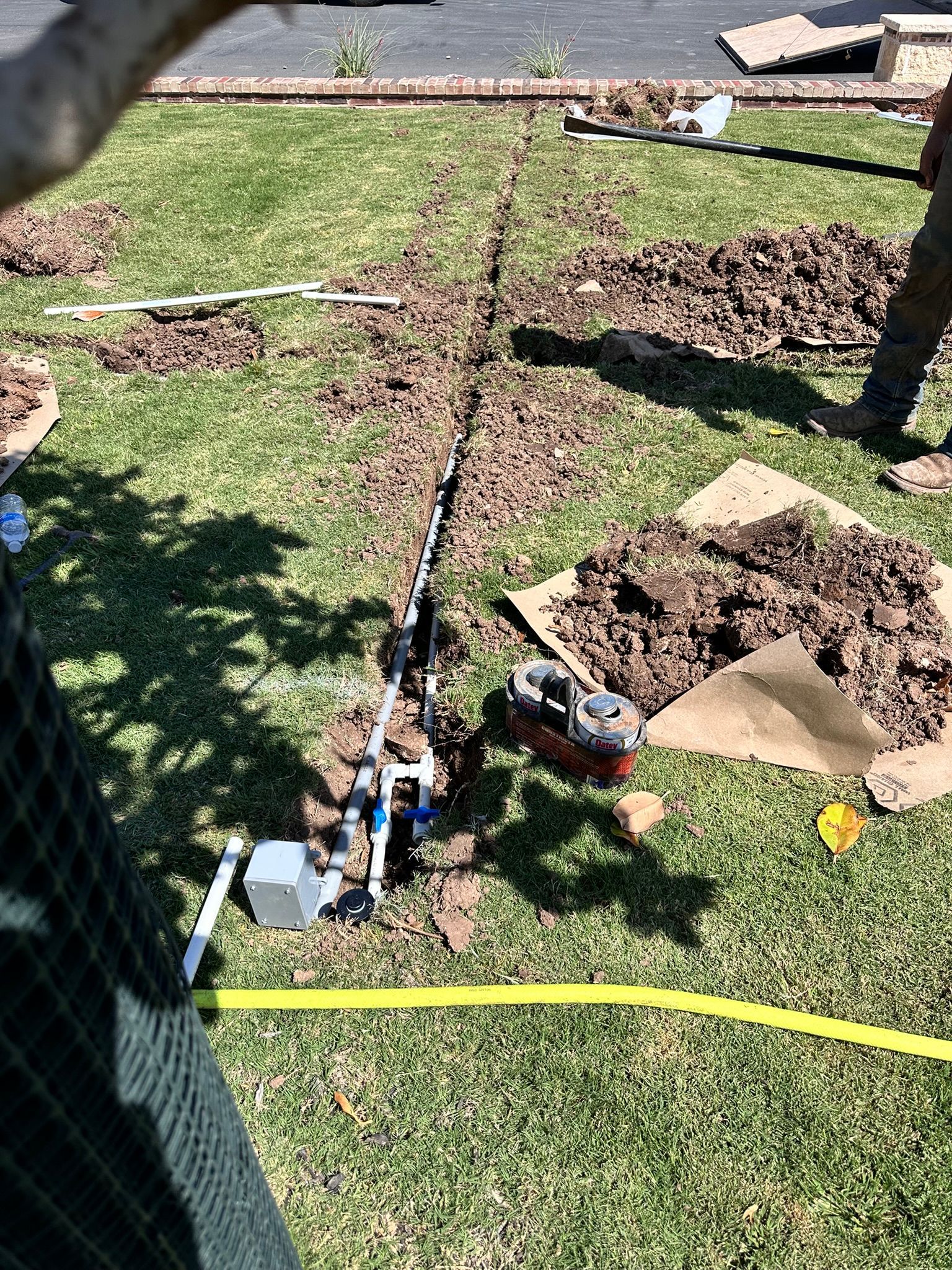 Trench dug in grass with irrigation system parts. Soil piles, yellow hose, blue valves, and a person’s legs are visible.
