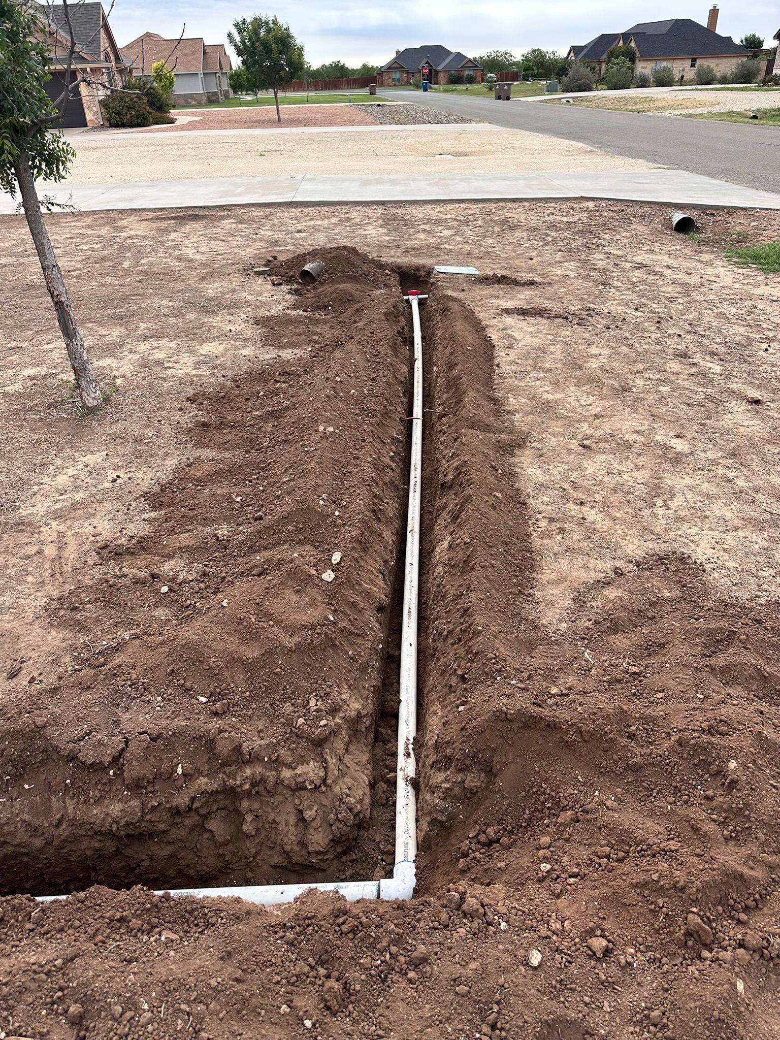 Trench with white PVC pipe installed, running through brown dirt. In a residential setting.