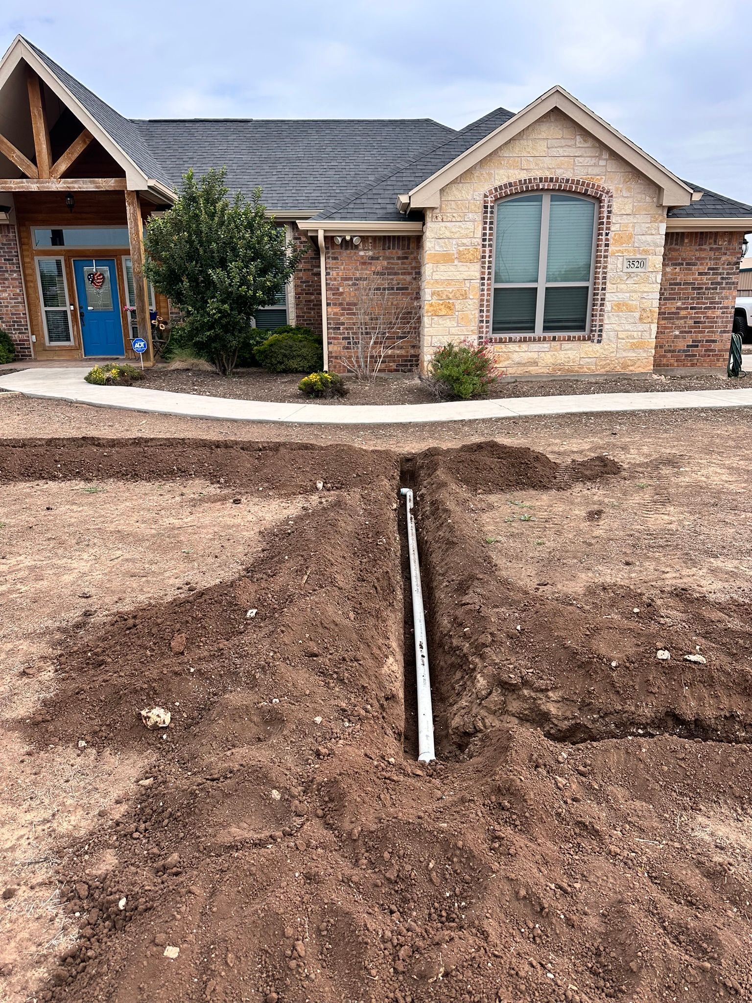 A trench dug in front yard with white pipe, leading to a brick and stone house.