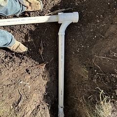 PVC pipes being installed in a trench in the dirt, next to a person's legs wearing jeans and boots.