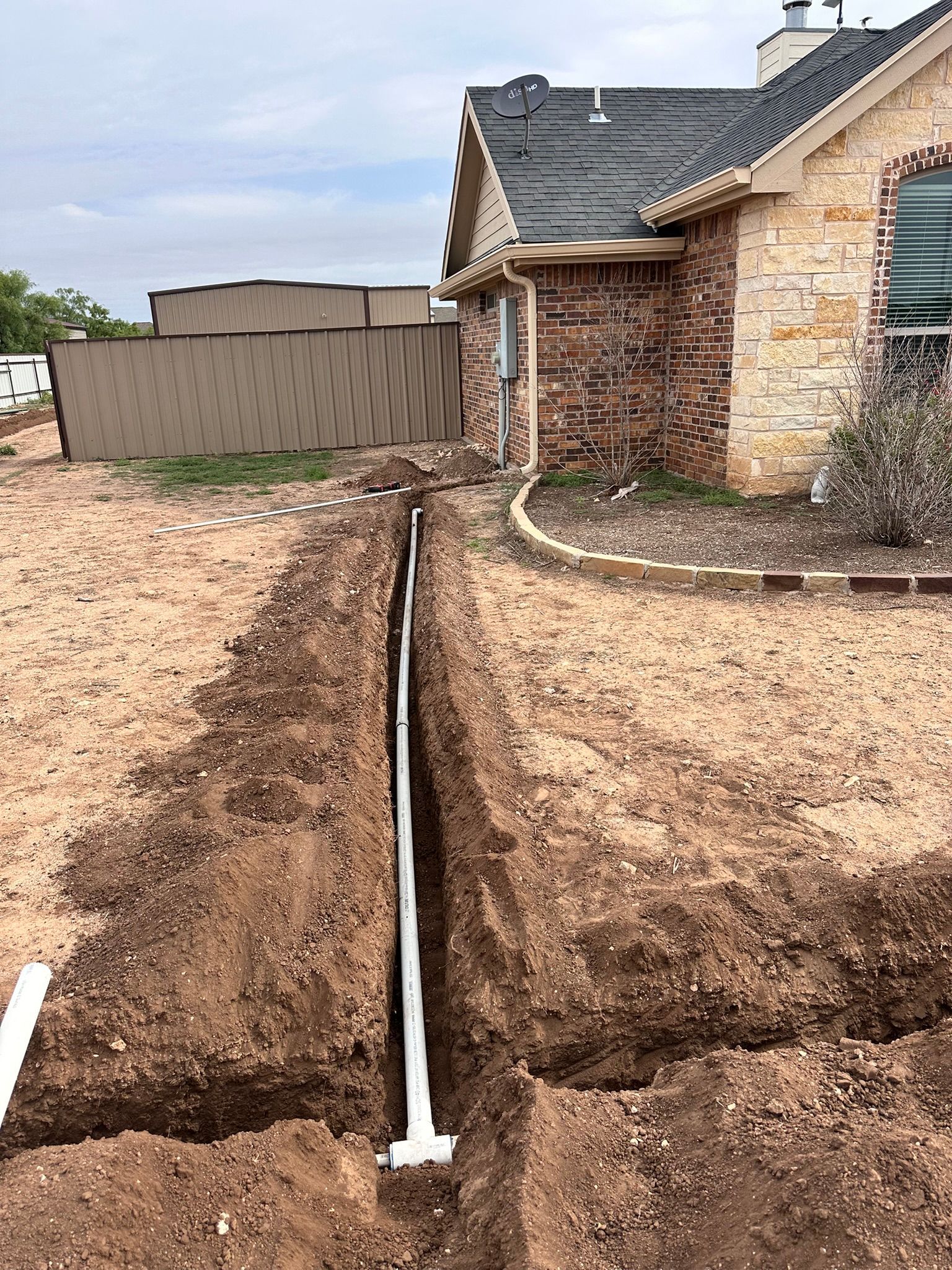 A trench in a yard with white PVC pipe running alongside a house. Brown soil and fence visible.