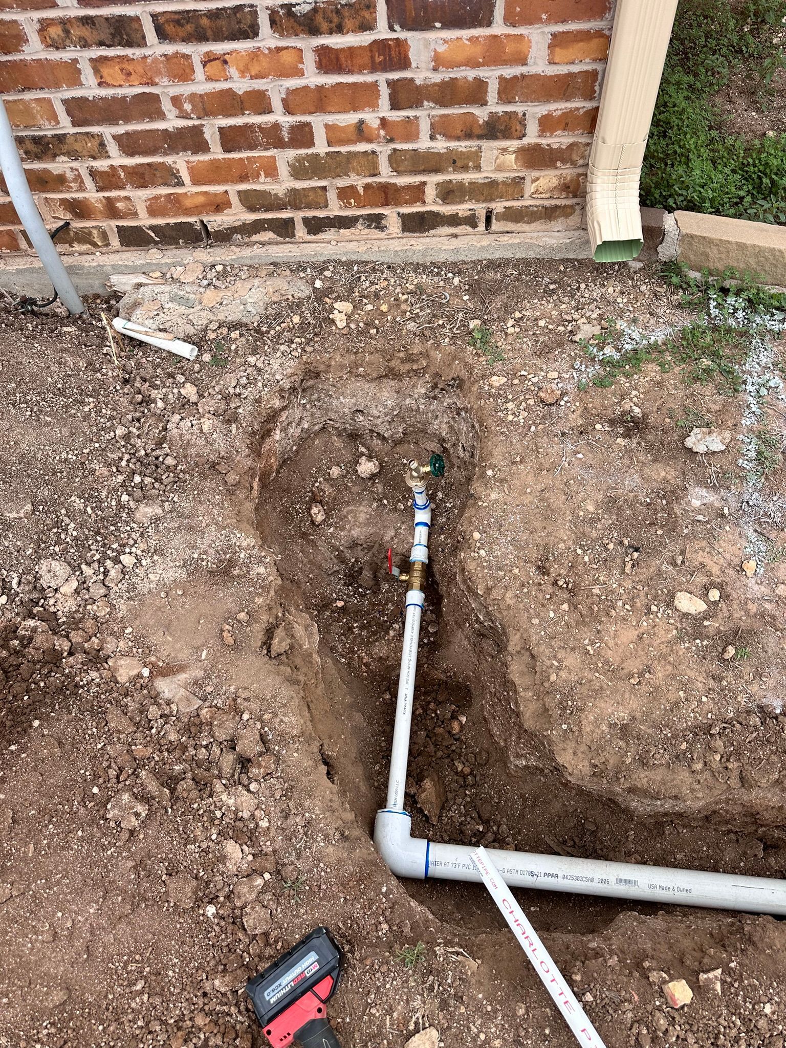 PVC pipe and water valve in a dirt trench near a brick wall.