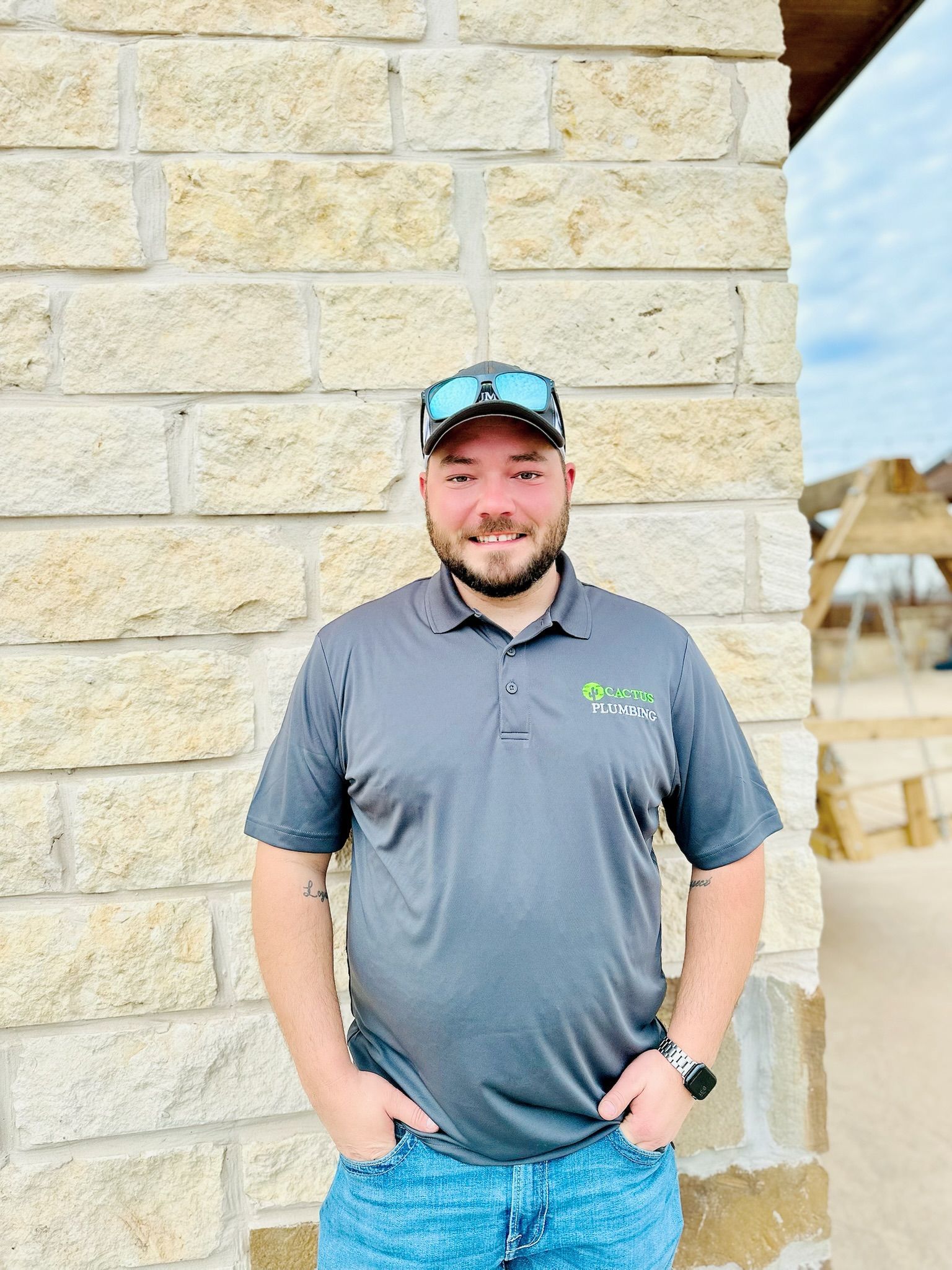 Man in gray polo shirt and cap, smiling, standing by stone wall; outdoor setting.