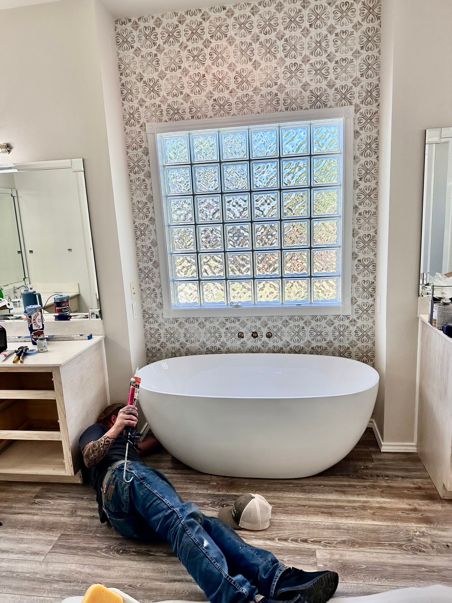 Man working on bathroom remodel, lying on floor. White tub, glass block window, and patterned wall.