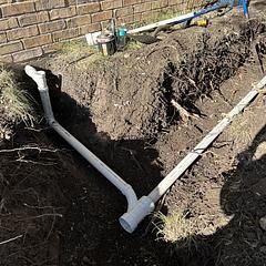 Gray PVC pipes in a trench alongside a brick wall; construction scene.