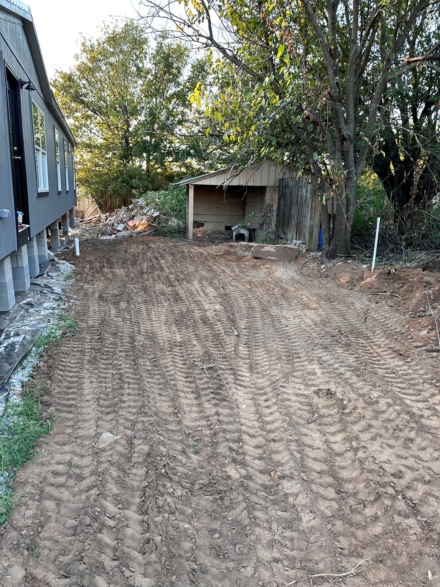 A dirt path with tire tracks leads towards a shed, beside a building with concrete pillars, trees overhead.
