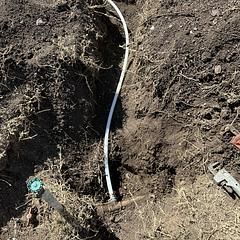 White pipe in a ditch connected to a blue and green faucet in dirt. A tool rests nearby.