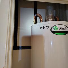 A white A.O. Smith water heater in a closet with pipes and date 9-9-13, surrounded by white and black walls.