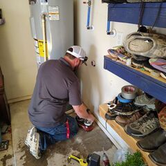 Plumber working on a pipe near a water heater in a garage, with shoes on shelves nearby.