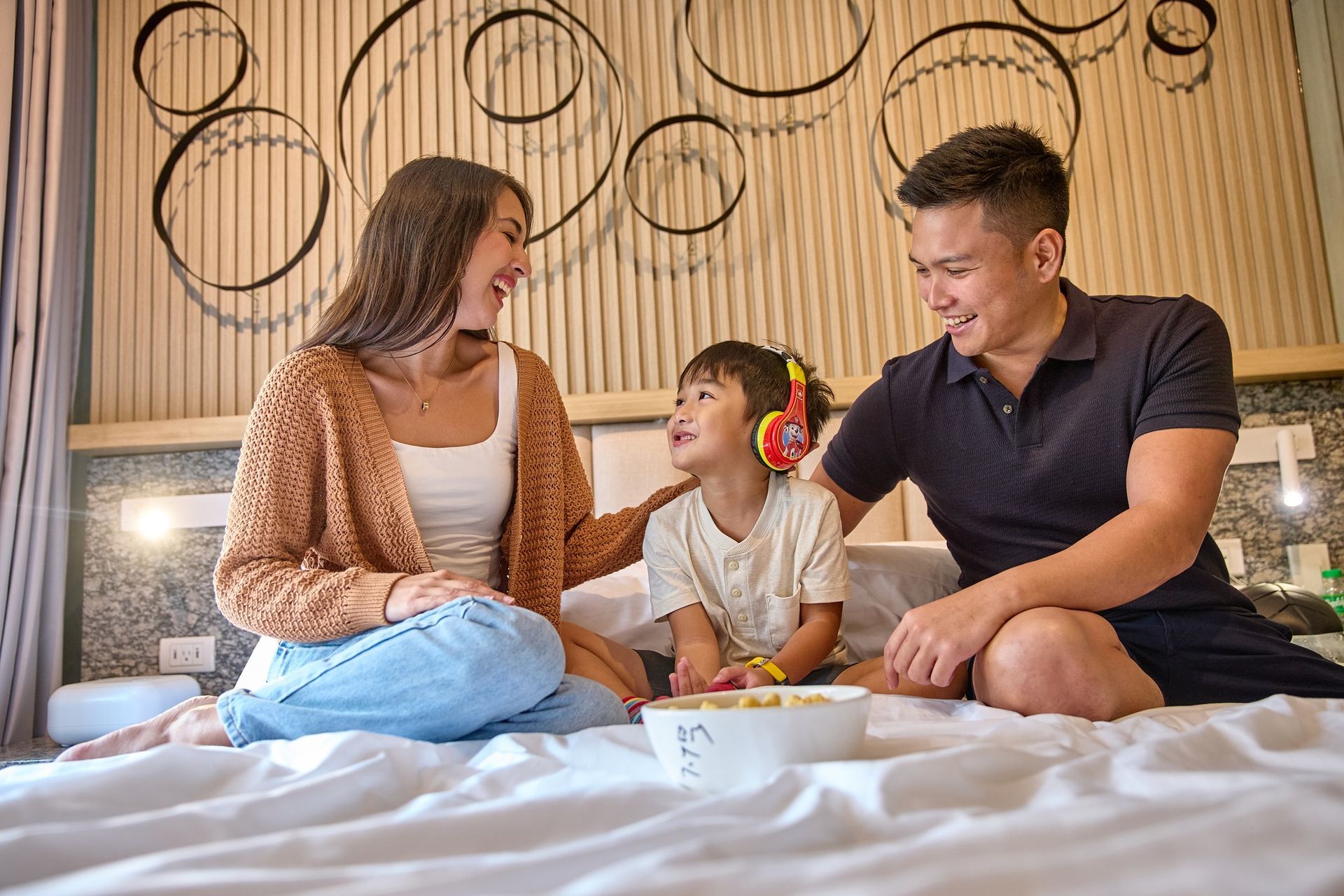 A family sits together on a white hotel bed, smiling and looking at a child wearing headphones.