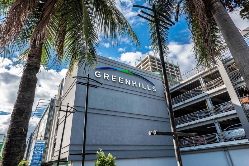 The Greenhills shopping center building with a sign under a bright blue sky, framed by palm trees and utility poles.