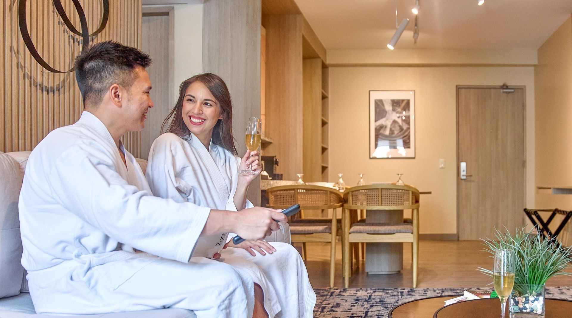 Couple in white robes on a sofa, smiling, holding drinks and remote, in a modern hotel room.