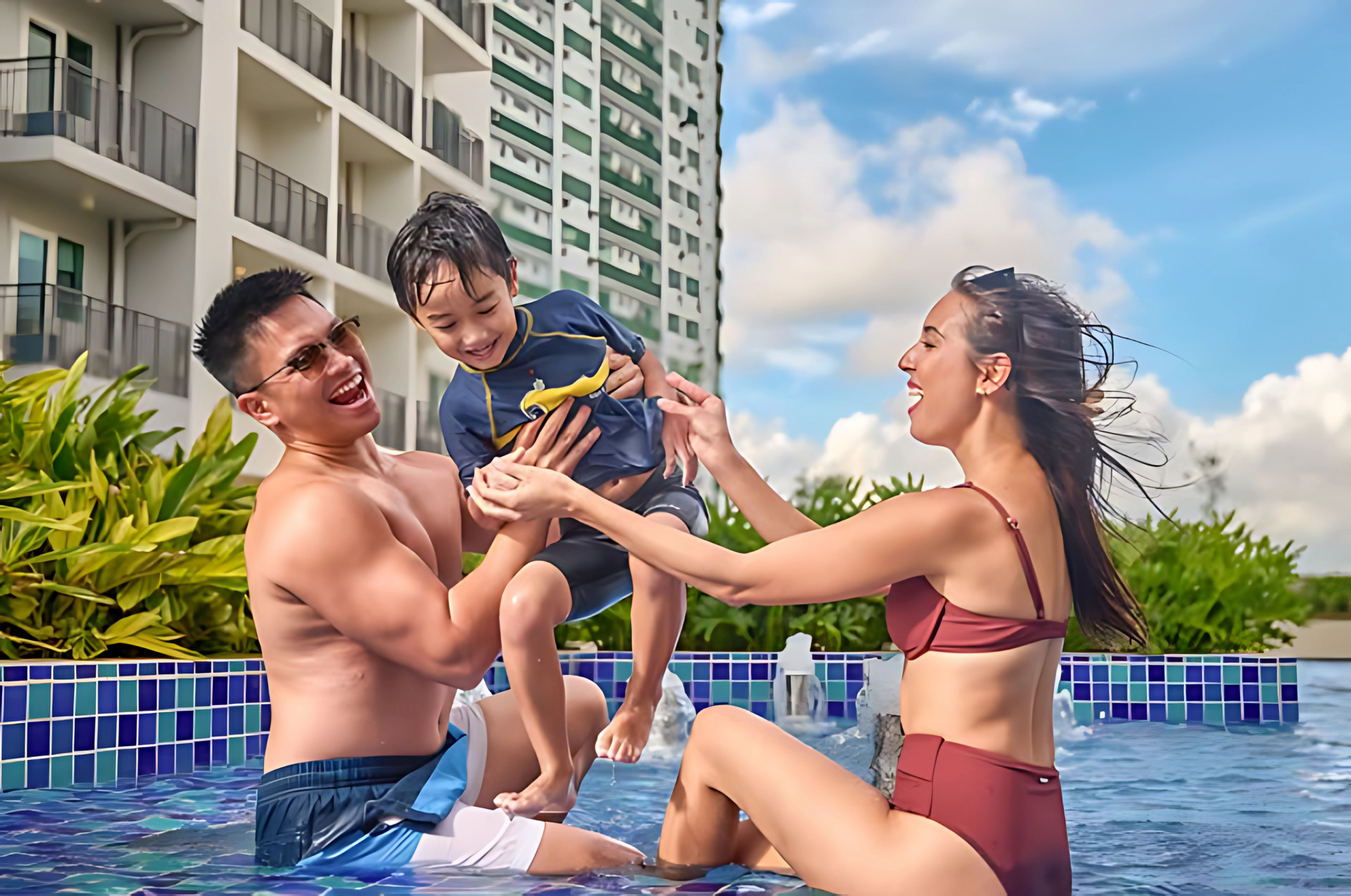 Family playing in a pool beside a hotel, with a child held between two adults under a blue sky.