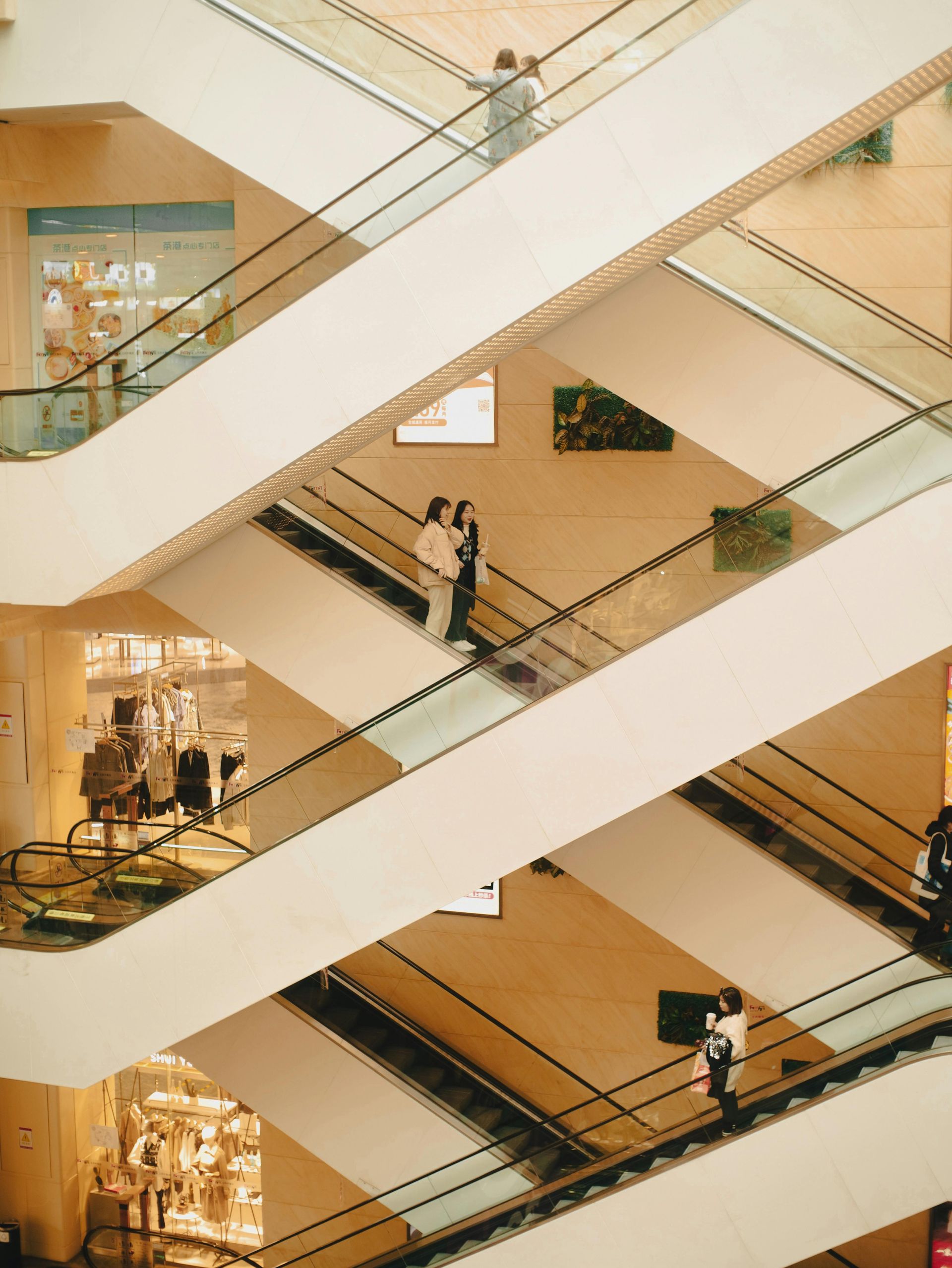 Escalators in a shopping mall, several people ride up and down. Beige and brown tones.