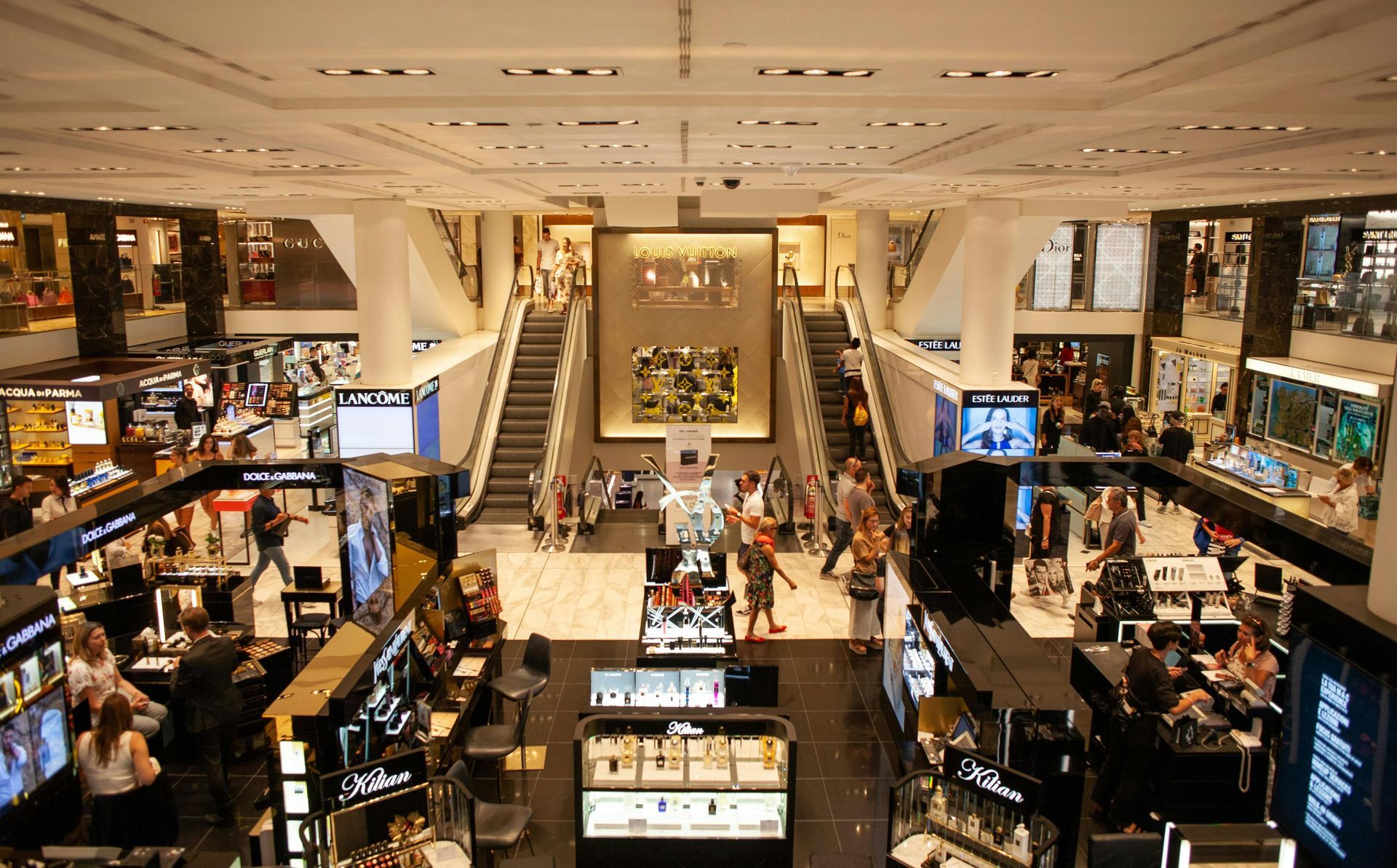 Multi-level department store interior with shoppers, escalators, and merchandise displays.