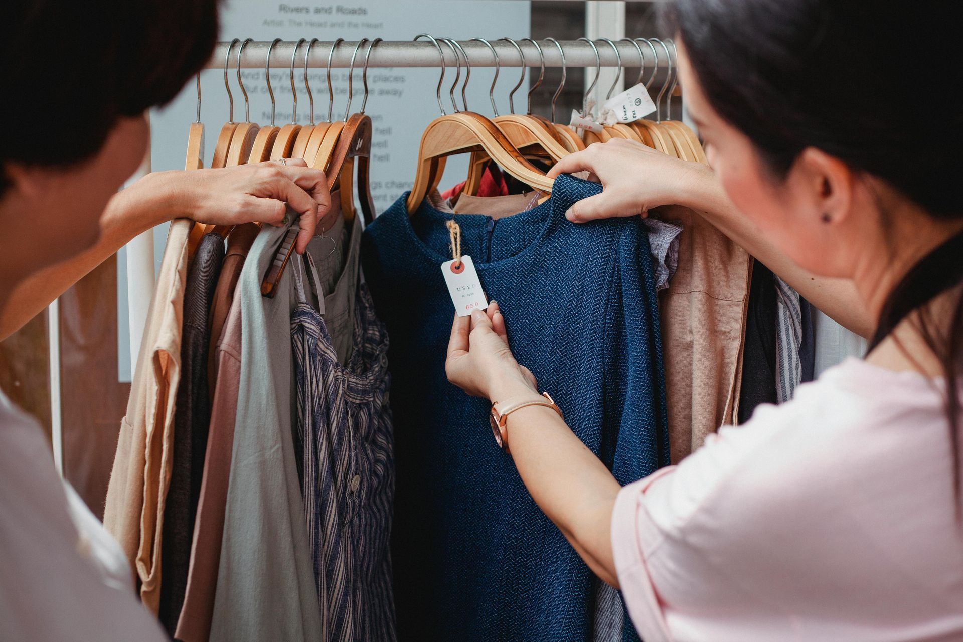 Two people examine clothes on a rack. One points at a blue dress with a price tag. Store setting.