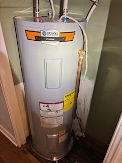 A gray State brand electric water heater standing in a utility closet with copper piping and white plumbing connections.