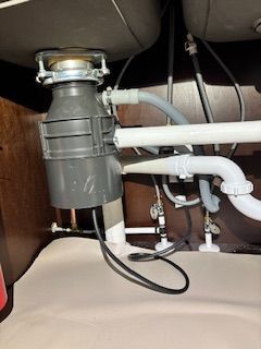 Under-sink view of a garbage disposal unit with connected drainage pipes and a power cord.