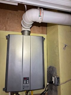 Wall-mounted tankless water heater with a white exhaust vent pipe, mounted on a yellow wall next to an electrical outlet.