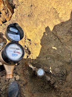 A person holds a water meter up to the camera with another meter partially buried in the dirt-filled utility hole below.