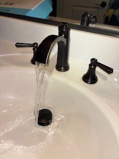 A chrome kitchen faucet sits on a dark stone countertop next to a circular stainless steel sink with marble-pattern walls.