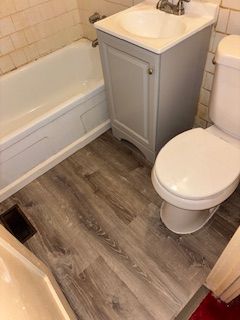A small bathroom with light gray vinyl flooring, a white bathtub, a matching gray vanity with a sink, and a white toilet.